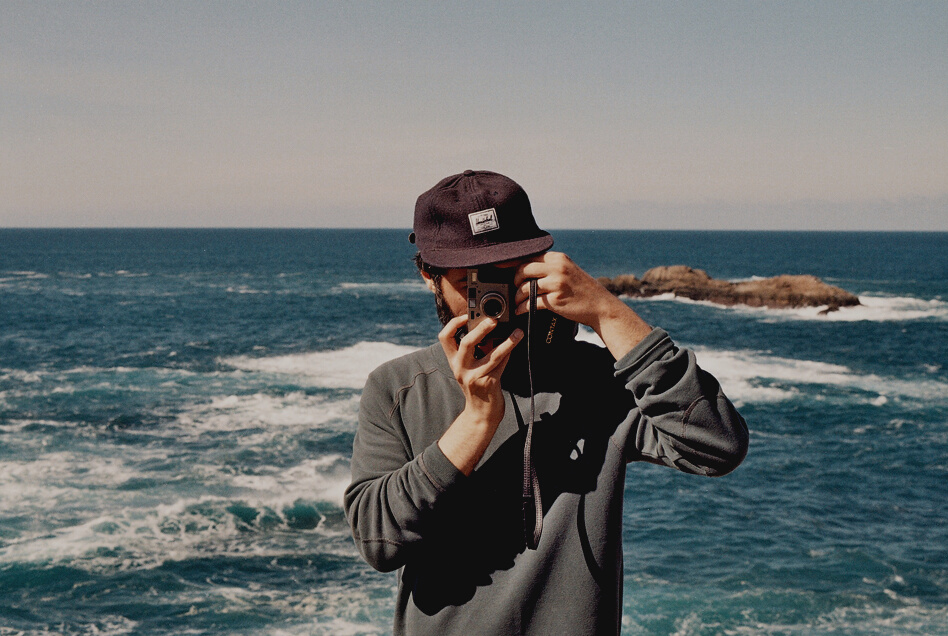 Man in Black Jacket Carrying Point and Shoot Camera in Front of Body of Water during Daytime